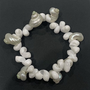 May include: A bracelet made of white seashells, arranged in a circular shape. The shells vary in size and shape, with some having a pearlescent sheen. The bracelet is set against a dark background, highlighting the natural beauty of the shells.
