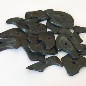 May include: A pile of dark gray, irregularly shaped buttons with a matte finish. Each button has a small hole in the center. The buttons are scattered on a white surface.