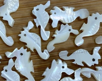 May include: A collection of iridescent white seahorse-shaped shell pieces with black dot eyes. The seahorses are scattered on a light brown wooden surface. These decorative shell pieces are ideal for crafting and jewelry making.