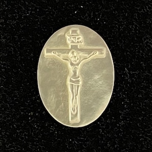 May include: Oval-shaped pendant with a carved image of Jesus on the cross. The pendant is a light, pearlescent color, and the details of the figure and cross are clearly defined. The background is a dark, textured surface.