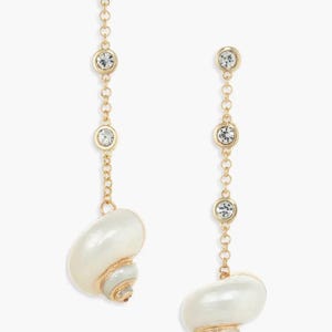 May include: A pair of gold-toned drop earrings. Each earring features three clear, round crystals connected by a delicate chain. The earrings end with a white seashell pendant with gold and gray accents. The earrings are designed for pierced ears.