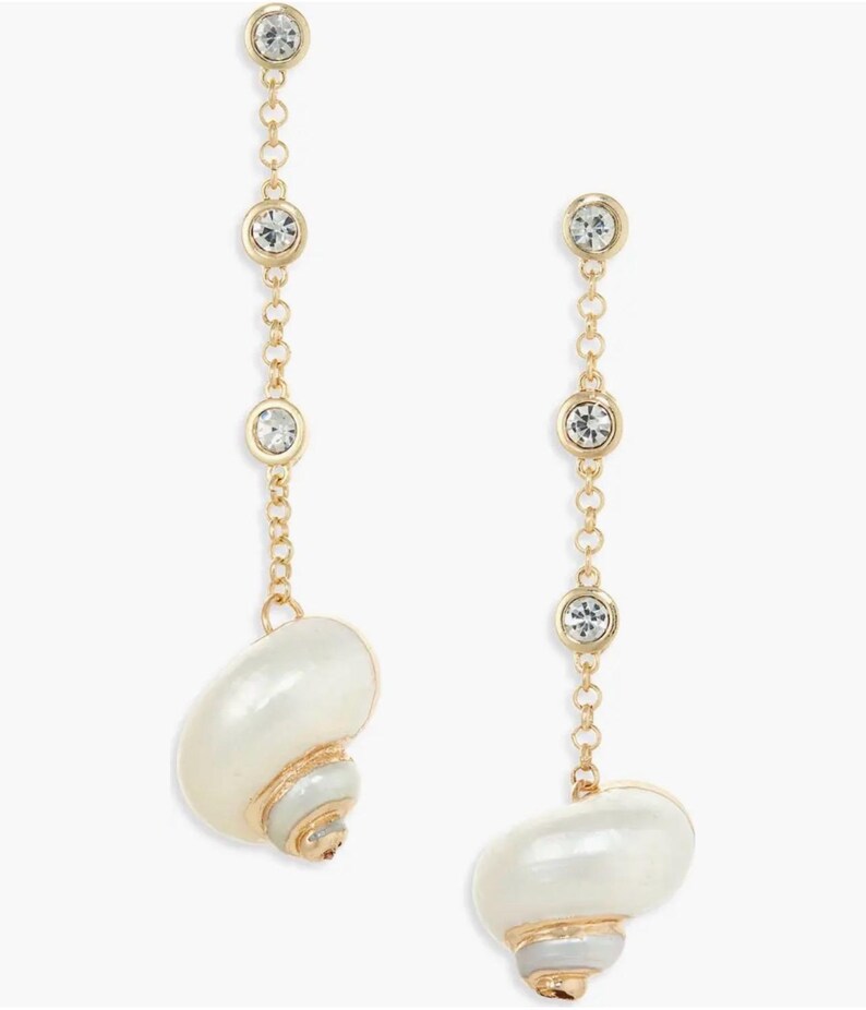 May include: A pair of gold-toned drop earrings. Each earring features three clear, round crystals connected by a delicate chain. The earrings end with a white seashell pendant with gold and gray accents. The earrings are designed for pierced ears.