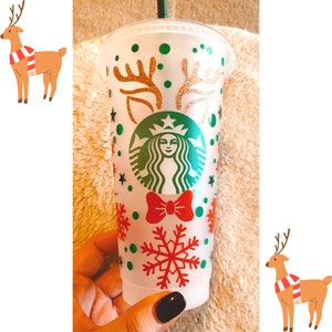 May include: A clear plastic Starbucks cold cup with a festive Christmas design. The cup features green and gold glitter accents, a red bow, and a large red snowflake. The Starbucks logo is in the center. The cup is decorated with reindeer antlers and green polka dots.