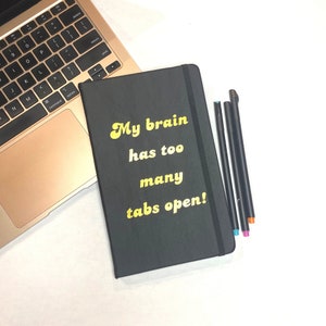 May include: A black notebook with the text "My brain has too many tabs open!" in yellow. Beside it are several black pens with coloured tips. A laptop is also visible. The background is white.