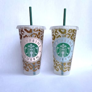 May include: Two clear plastic tumblers with lids and green straws. The tumblers have a leopard print design and the text "CAFFEINE QUEEN" with a green logo. One tumbler has a pink background, the other has a light blue background.
