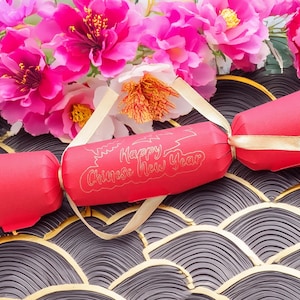 Red Chinese New Year cracker with gold foil “Happy Chinese New Year” design, tied with gold satin ribbon, styled with bright pink blossom flowers on a patterned black and gold background.
