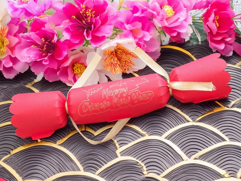 Red Chinese New Year cracker with gold foil “Happy Chinese New Year” design, tied with gold satin ribbon, styled with bright pink blossom flowers on a patterned black and gold background.