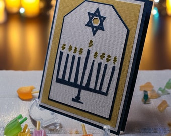 May include: A white card with a blue menorah and a gold Star of David on a gold background. The card is surrounded by a blue border. The card is sitting on a white surface with colorful dreidels.