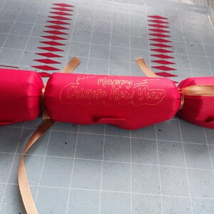 Red cracker with gold foil “Happy Chinese New Year” message, tied with gold ribbon, placed on a craft mat background alongside paper cut-outs.