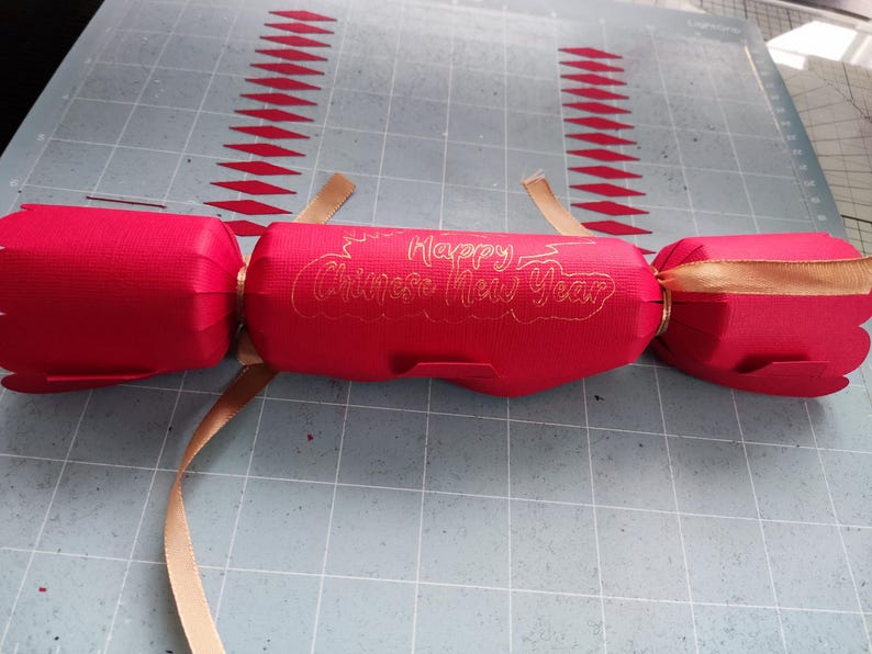 Red cracker with gold foil “Happy Chinese New Year” message, tied with gold ribbon, placed on a craft mat background alongside paper cut-outs.