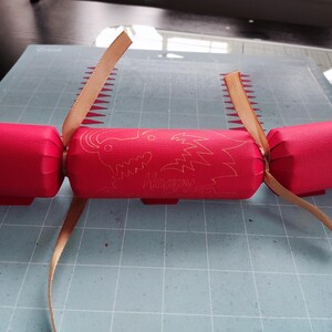 Handmade red Chinese New Year cracker with gold foil dragon design, tied with gold ribbon, shown flat on a crafting mat during assembly.