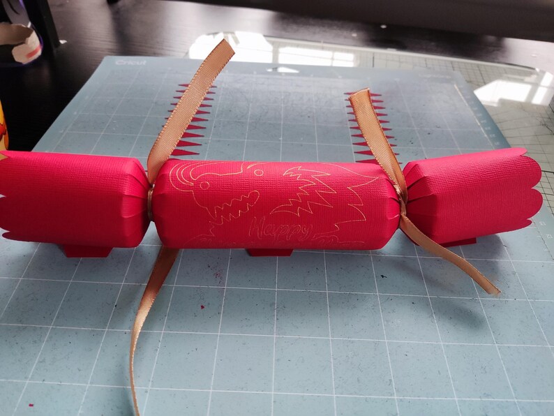Handmade red Chinese New Year cracker with gold foil dragon design, tied with gold ribbon, shown flat on a crafting mat during assembly.