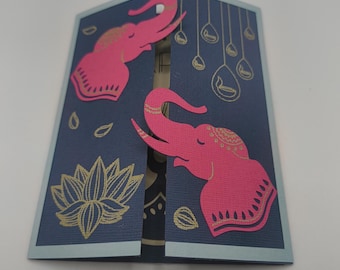 Op de afbeelding: Een marineblauwe wenskaart met een lichtblauwe rand, met roze olifantenuitsparingen en gouden accenten. De kaart is versierd met een lotusbloem, druppelvormen en andere decoratieve elementen. De kaart is open en onthult een patroon aan de binnenkant.