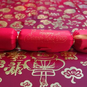 Red cracker with gold foil “Happy Chinese New Year” lettering, tied with gold ribbon, displayed on a red and gold patterned festive backdrop with traditional Chinese motifs.