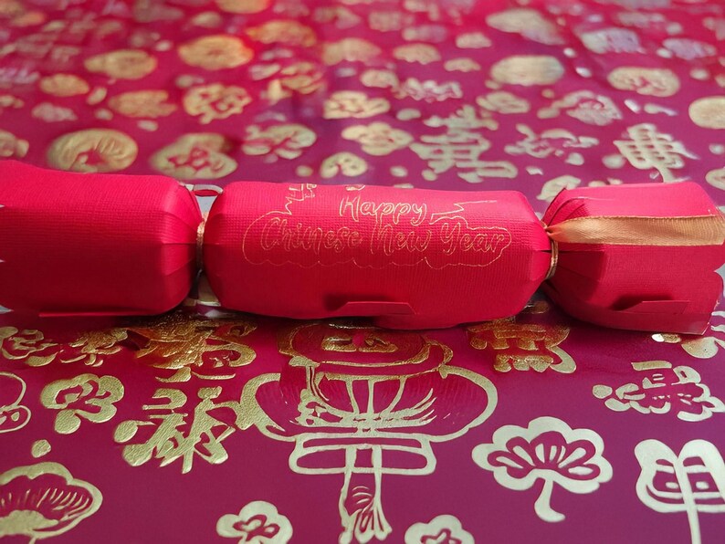 Red cracker with gold foil “Happy Chinese New Year” lettering, tied with gold ribbon, displayed on a red and gold patterned festive backdrop with traditional Chinese motifs.