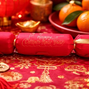 Bright Chinese New Year lifestyle photo showing a red personalised party cracker with gold lettering, styled on a red and gold festive table with mandarins, gold ingots, coins, and lanterns in the background.