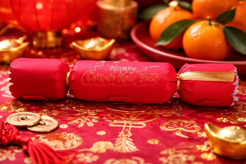 Bright Chinese New Year lifestyle photo showing a red personalised party cracker with gold lettering, styled on a red and gold festive table with mandarins, gold ingots, coins, and lanterns in the background.