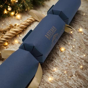 May include: A navy blue Christmas cracker with gold detailing and a gold ribbon. The word "BARBARA" is printed in gold. The cracker rests on a fluffy, light brown surface, with Christmas lights and a Christmas tree visible in the background.
