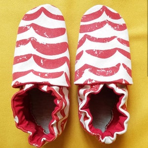 May include: A pair of baby booties with a red and white wave pattern. The booties have a red interior and a white exterior with a repeating red wave design. The soles are soft and flexible.