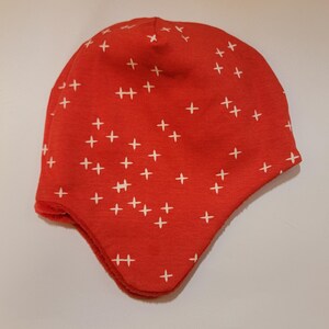 May include: Red fleece hat with a white plus sign pattern. The hat has a fold-down ear flap.