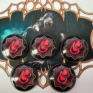 May include: Five circular buttons featuring a black background with a red eight-pointed star and a red double-drop symbol. The buttons are arranged in a cluster over a decorative backing card that displays a dark, illustrated figure.