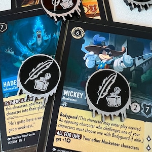 May include: Two black and silver tokens with a quill and inkwell design. The tokens are from the Disney Lorcana Trading Card Game. The text on the card reads "Bodyguard (This character may enter play exerted. An opposing character who challenges one of your characters must choose one with Bodyguard if able.) ALL FOR ONE Your other Musketeer characters get +10".