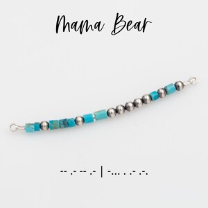 May include: A curved necklace with turquoise and silver beads. The necklace has the words "Mama Bear" in black script above it. Below the necklace is a line of Morse code.
