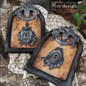May include: Two ornate black framed wall decor pieces with a brown background. Each piece features a silver keyhole with a silver key and decorative silver embellishments.