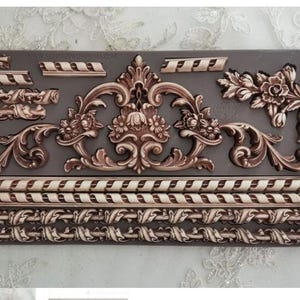 May include: A decorative mold featuring various ornate, vintage-style designs. The designs include floral elements, scrolls, and borders in shades of brown and cream, set against a dark gray background. The mold is rectangular and likely used for crafting or decorative purposes.