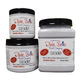May include: Three jars of Dixie Belle Paint Company BOSS paint. The jars are labelled with the company name, product name, and size. The smallest jar is 8 oz, the middle jar is 16 oz, and the largest jar is 32 oz. The paint is grey.