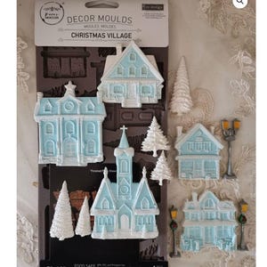 May include: A Christmas village mould featuring miniature houses, trees, and street lamps. The houses are light blue with white detailing. The mould measures 12.7cm by 25.4cm.