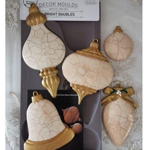 May include: A set of decorative molds for making bright baubles. The molds are in various shapes, including teardrop, bell, and round, with a cream-colored base and gold accents. The package is 5" x 10".