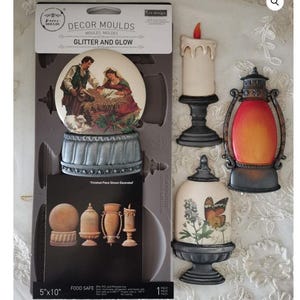 May include: A collection of decorative molds and finished pieces. The molds include a nativity scene, a candle, a lantern, and a cloche with a butterfly design. The text on the packaging reads "Glitter and Glow".
