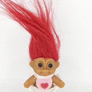 Puede incluir: Un pequeño muñeco troll de pelo rojo, con un pañal blanco y un babero rosa con un corazón rojo. El muñeco tiene ojos marrones y una cara sonriente.