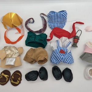 Puede incluir: Una colección de ropa y accesorios para muñecas, que incluye un vestido a rayas, una camisa a rayas rojas y blancas, un chaleco verde, unos pantalones cortos marrones, un chaleco dorado y plateado, un par de sandalias marrones, un par de zapatos negros, un vestido rosa con un diseño de perro, una linterna, una cinta rosa, una corona dorada y un sombrero a rayas.