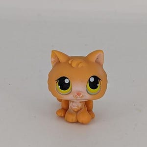 May include: A small, orange cat figurine with large black eyes and yellow irises. The toy cat has a white muzzle and paws, and is in a sitting position. The cat's fur is a solid orange color.