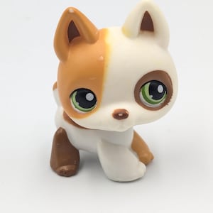 May include: A small plastic dog figurine with a white base coat, featuring a large brown patch covering the right ear and side of the head. The figure has oversized green eyes with black pupils, a small brown nose, and brown markings on its legs.