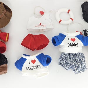 Puede incluir: Una colección de ropa de muñeca, que incluye un sombrero de vaquero marrón, un uniforme rojo y dorado, un atuendo de salvavidas, una camiseta "I love Daddy", un uniforme de árbitro y otros accesorios. Los atuendos son de varios colores y estilos.