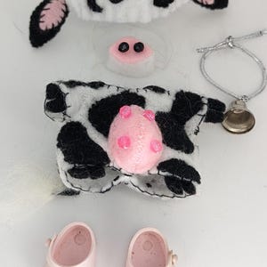 Puede incluir: Un disfraz de vaca para una muñeca, que incluye un vestido con lunares negros y blancos, un sombrero de vaca con una flor amarilla, una nariz rosa y zapatos rosas.