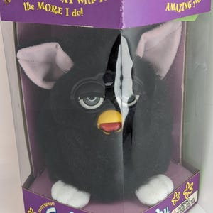 Könnte beinhalten: Ein schwarzes Furby-Spielzeug in seiner Originalverpackung. Das Furby hat einen schwarzen Körper, rosa Ohren, weiße Füße und einen gelben Schnabel. Die Verpackung ist lila und trägt die Aufschrift "Furby" und "Electronic".