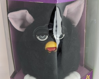 May include: A black Furby toy in its original packaging. The Furby has a black body, pink ears, white feet, and a yellow beak. The packaging is purple and has the words "Furby" and "Electronic" printed on it.