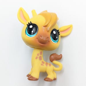May include: A yellow giraffe figurine with brown spots and a brown tail. The giraffe has large black eyes with blue accents and a smiling expression. The ears and mane are a darker shade of yellow.