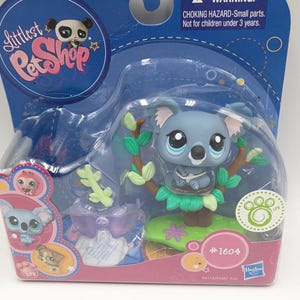 May include: A Littlest Pet Shop toy featuring a blue koala figure with large eyes, perched on a tree branch. The toy is packaged in a clear plastic blister pack with a blue backing, and the package includes a warning about small parts.