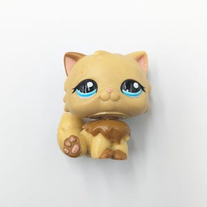 May include: A tan-colored cat figurine with large blue eyes and brown accents. The toy cat has a detailed design, including a pink nose and brown paws. The cat is sitting with its tail curled up, and the head is slightly turned.