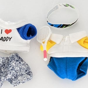 Puede incluir: Cuatro conjuntos de peluche para juguetes con diferentes diseños. Los conjuntos incluyen una camiseta amarilla y azul con la frase "I love Grandpa" impresa, una camiseta azul y blanca con la frase "I love Daddy" impresa, una camiseta morada y blanca con la frase "I love my Sister" impresa y una camiseta blanca y amarilla con una raqueta de tenis.