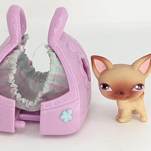 Op de afbeelding: Een roze en wit gestreepte dieren draagtas met een bruin en tan chihuahua beeldje. De draagtas heeft een bloemenontwerp en opent om een bed binnenin te onthullen.