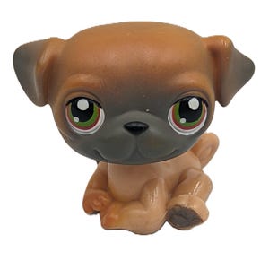 Puede incluir: Una pequeña figura de perro pug de color canela con cabeza marrón y hocico gris. El pug tiene ojos grandes y expresivos con iris verdes y pupilas negras. La figura está en posición sentada, con las patas extendidas.