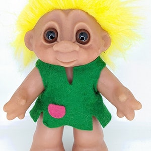 May include: A small, yellow-haired troll doll wearing a green dress with a pink patch. The doll has brown eyes and a friendly smile.
