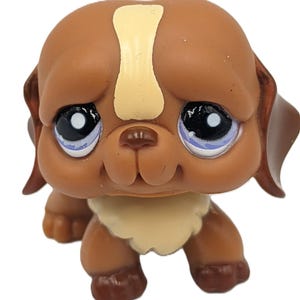 May include: A brown and tan puppy figurine with large, expressive blue eyes. The toy dog has a cream-colored patch on its forehead and a cream-colored chest. The puppy is in a sitting position and has brown floppy ears.