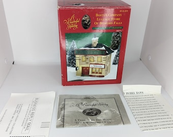 May include: A red box with a white and gold building on the front. The box is labeled "Boston Company Luggage Store of Bedford Falls" and "A Wonderful Holiday, A Tribute to the Movie "It's a Wonderful Life".